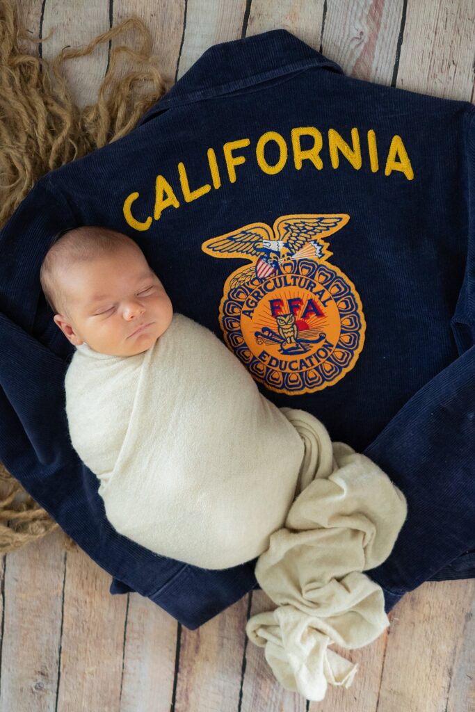Newborn James lies on a soft gray blanket, draped with his parents’ FFA jacket, highlighting their shared history. The natural tones and muted lighting create a timeless feel in this Sacramento, CA newborn photography session.